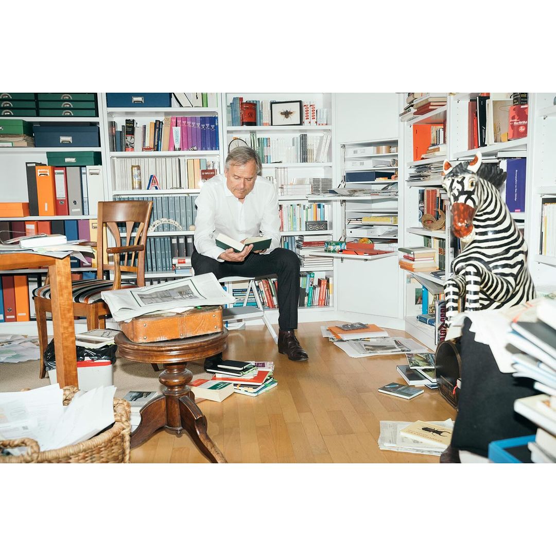 Man sitting and reading in a book-filled room with a zebra statue. The room has bookshelves, papers, and a table.