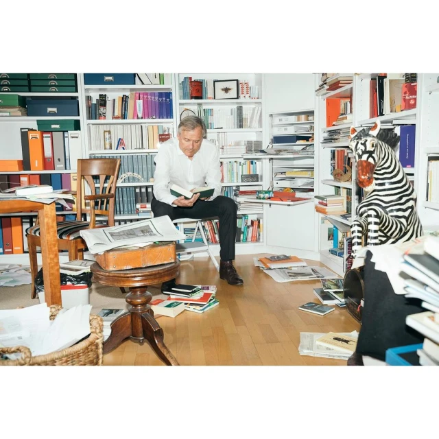 Man sitting and reading in a book-filled room with a zebra statue. The room has bookshelves, papers, and a table.