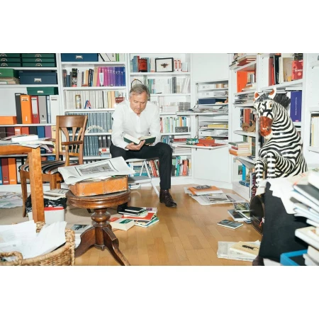 Man sitting and reading in a book-filled room with a zebra statue. The room has bookshelves, papers, and a table.