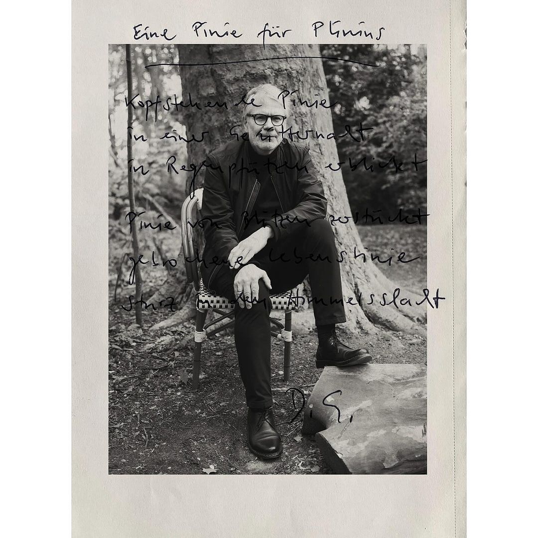 Black and white photo of an adult male with glasses and a jacket, sitting on a chair outdoors with trees in the blurred background.