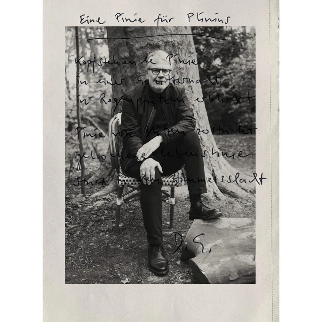 Black and white photo of an adult male with glasses and a jacket, sitting on a chair outdoors with trees in the blurred background.