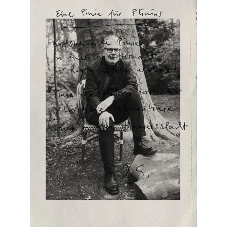 Black and white photo of an adult male with glasses and a jacket, sitting on a chair outdoors with trees in the blurred background.