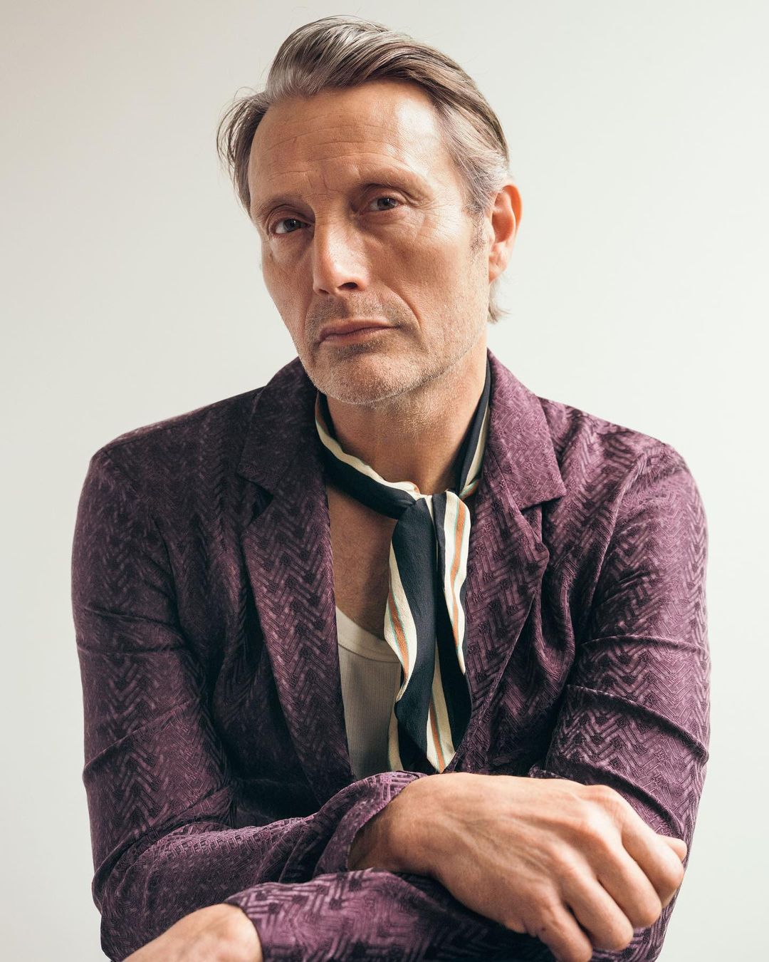 Portrait of Mads Mikkelsen wearing a patterned suit jacket and a loosely tied neck scarf, posing indoors against a neutral backdrop.