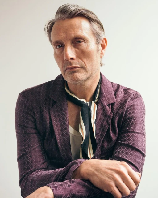 Portrait of Mads Mikkelsen wearing a patterned suit jacket and a loosely tied neck scarf, posing indoors against a neutral backdrop.