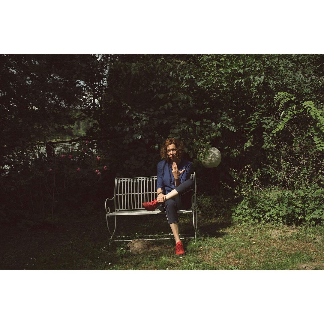Adult woman sits cross-legged on a wrought-iron bench in an outdoor garden setting, wearing blue jacket and red shoes.