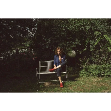 Adult woman sits cross-legged on a wrought-iron bench in an outdoor garden setting, wearing blue jacket and red shoes.