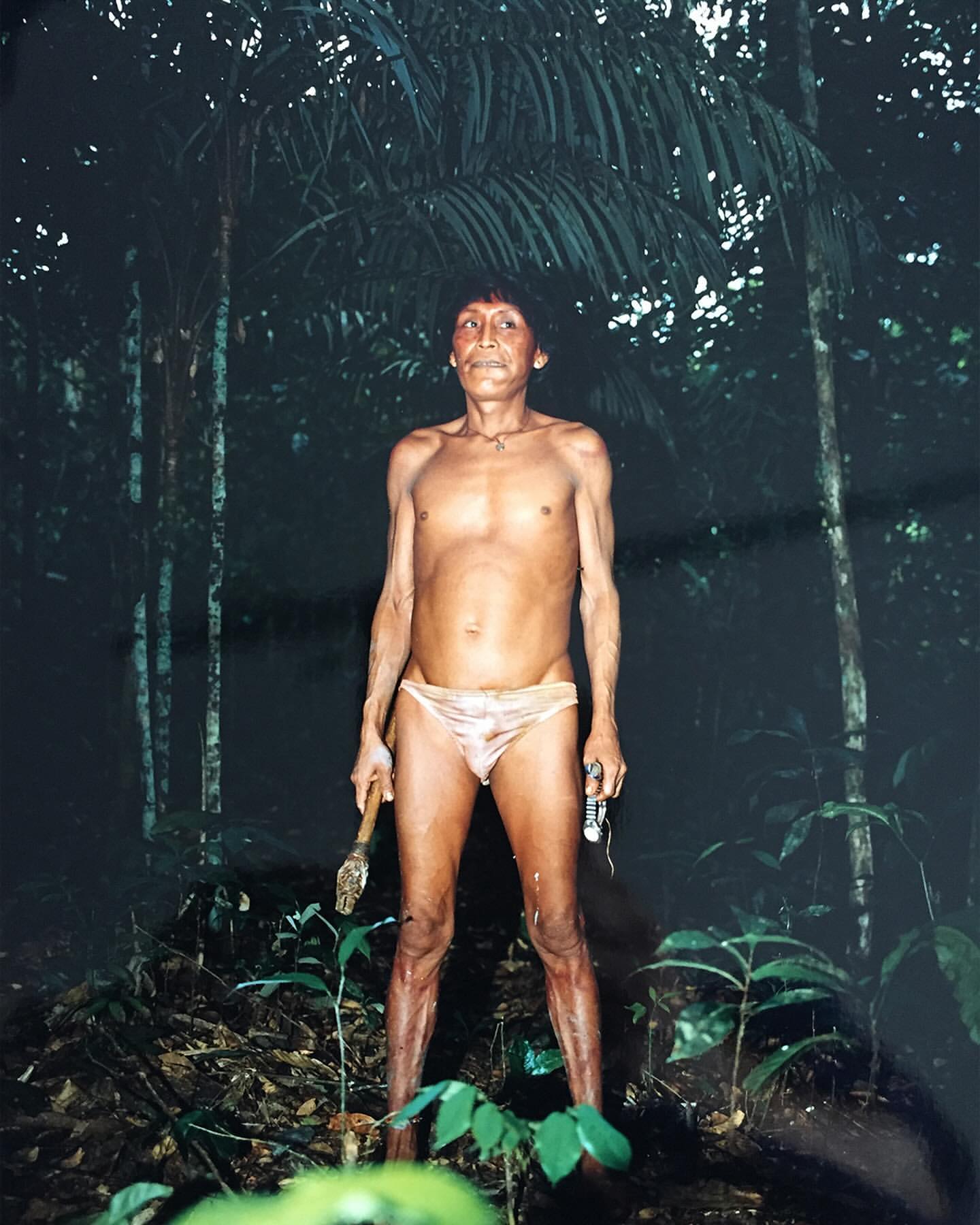 Bare-chested Indigenous man stands in a jungle environment, holding an axe and flashlight. Lush green plants and trees surround him.