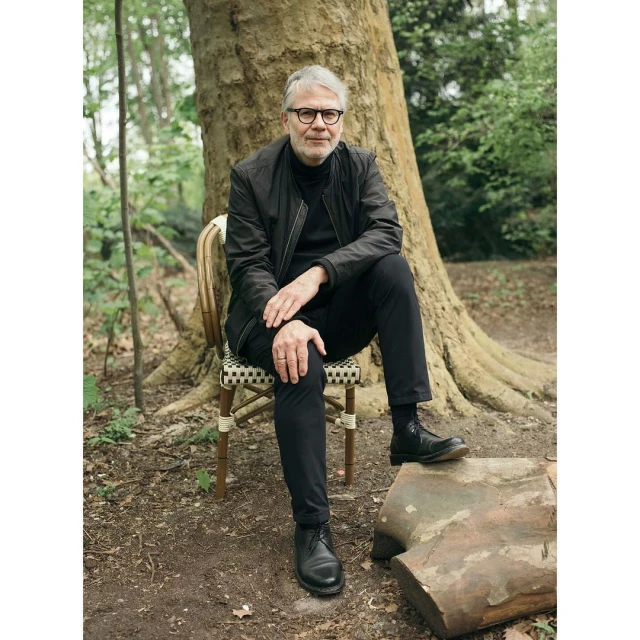 Mature adult man sitting on chair outdoors in wooded area, wearing a black jacket, pants, and boots with a tree trunk behind him.