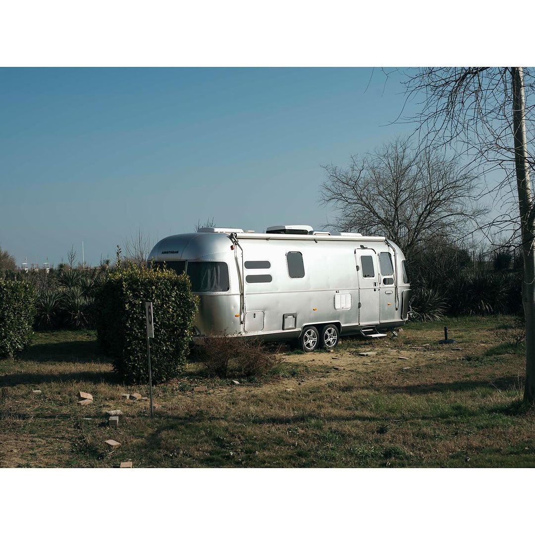 Silver Airstream trailer parked on a grassy lot with trees and bushes under a clear blue sky, creating a peaceful scene.