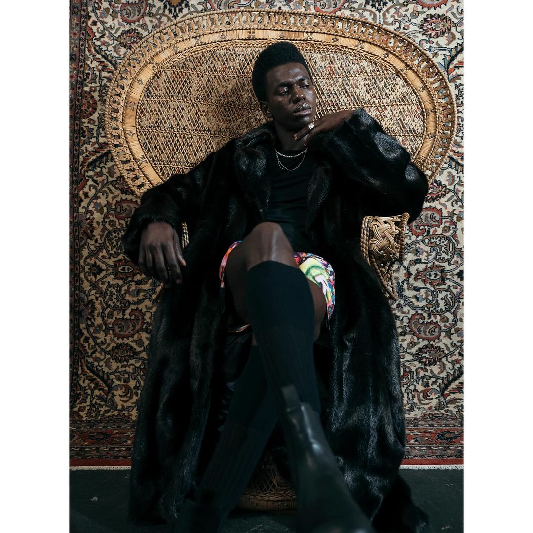 A dark-skinned man wearing a long, dark coat poses seated in a wicker chair against an ornate patterned rug indoors.