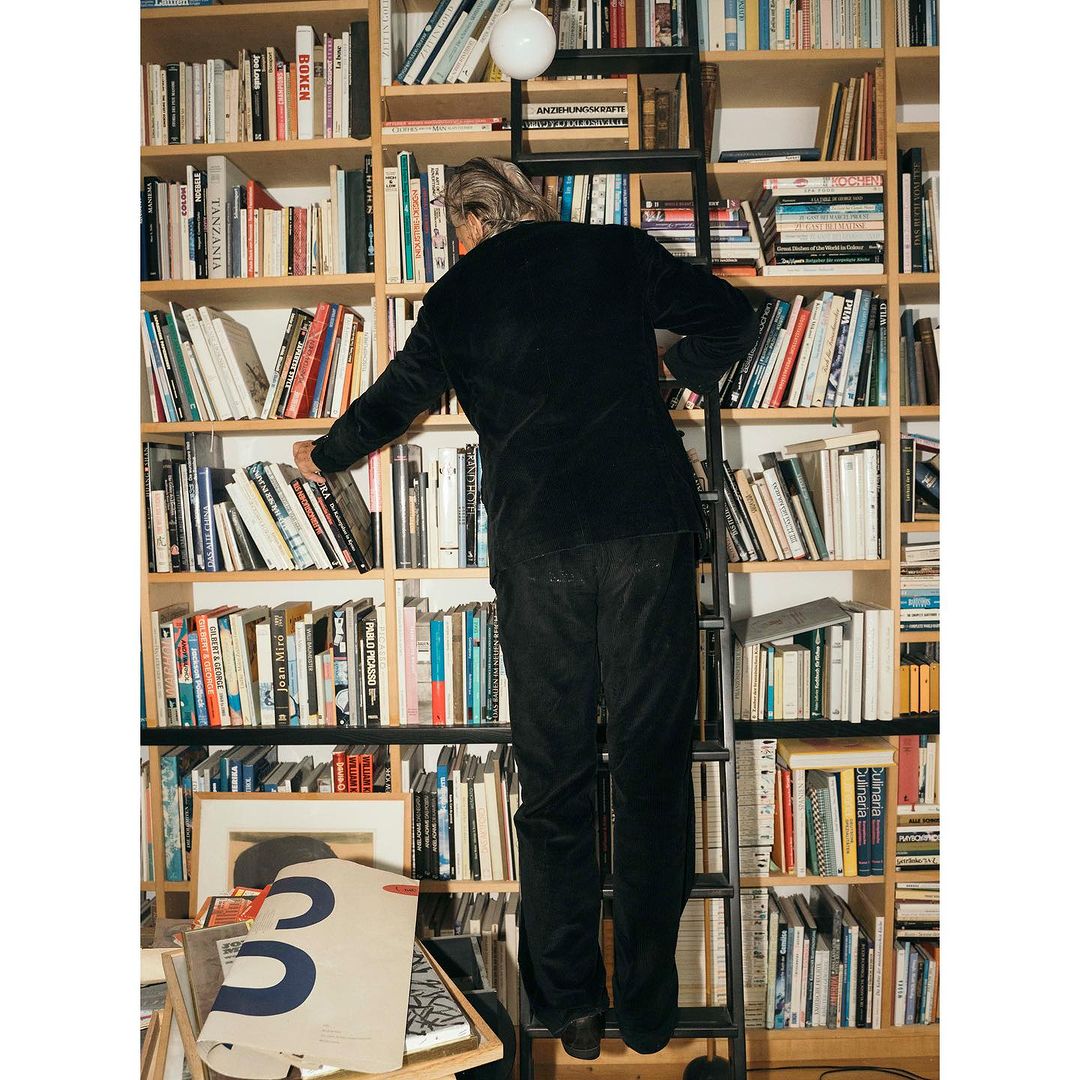 Man on a ladder reaching for a book on a shelf, indoors, in a room with a large bookcase, shot from a low angle looking up.