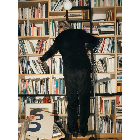 Man on a ladder reaching for a book on a shelf, indoors, in a room with a large bookcase, shot from a low angle looking up.