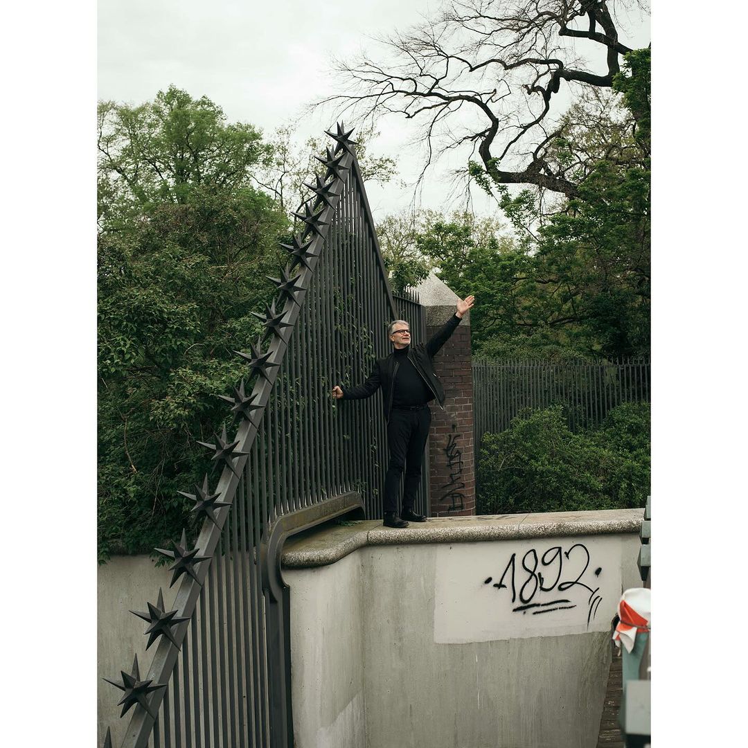 Full shot of adult man standing in front of a metal gate with star details, raising his arms. The background is a green outdoor park setting.