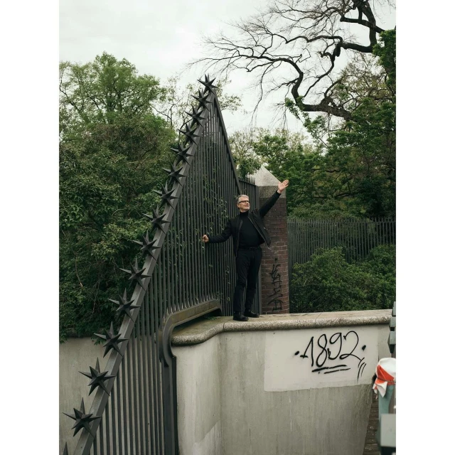 Full shot of adult man standing in front of a metal gate with star details, raising his arms. The background is a green outdoor park setting.