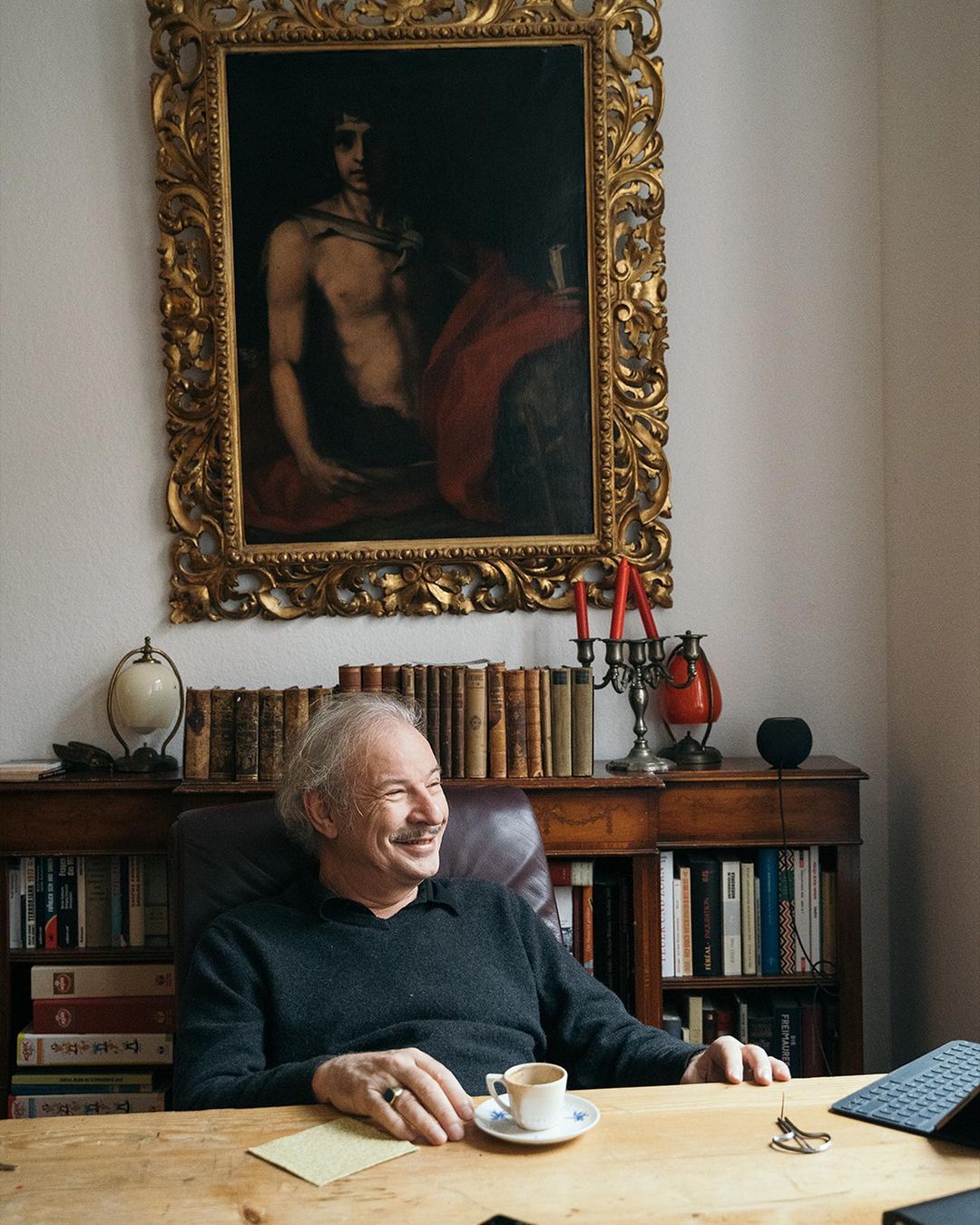 Indoor shot of a smiling mature man with white hair sitting at a desk with books, a painting, and a cup of coffee.