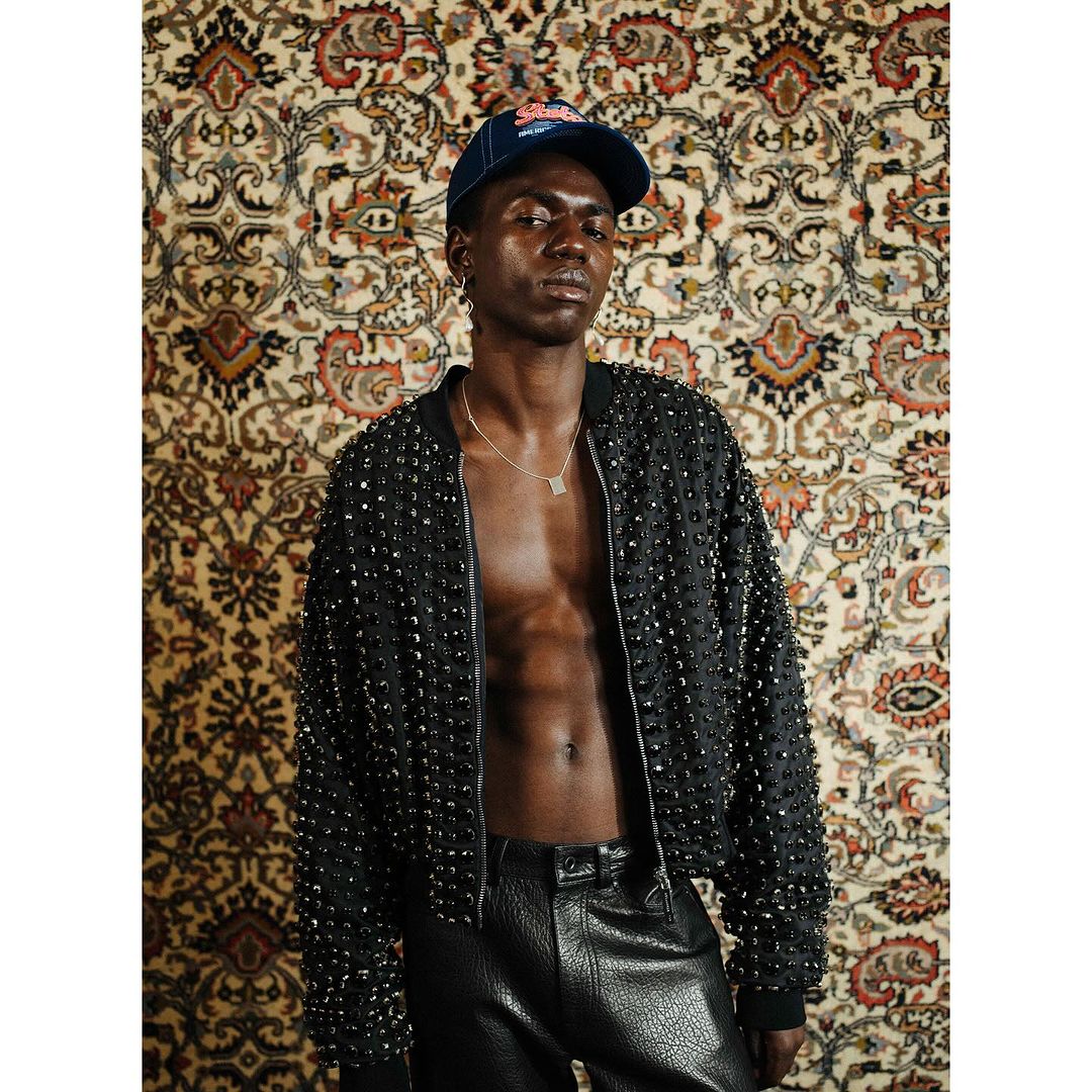 An African male model poses in black studded jacket and leather pants, accessorized with a necklace and baseball cap, against patterned wallpaper.