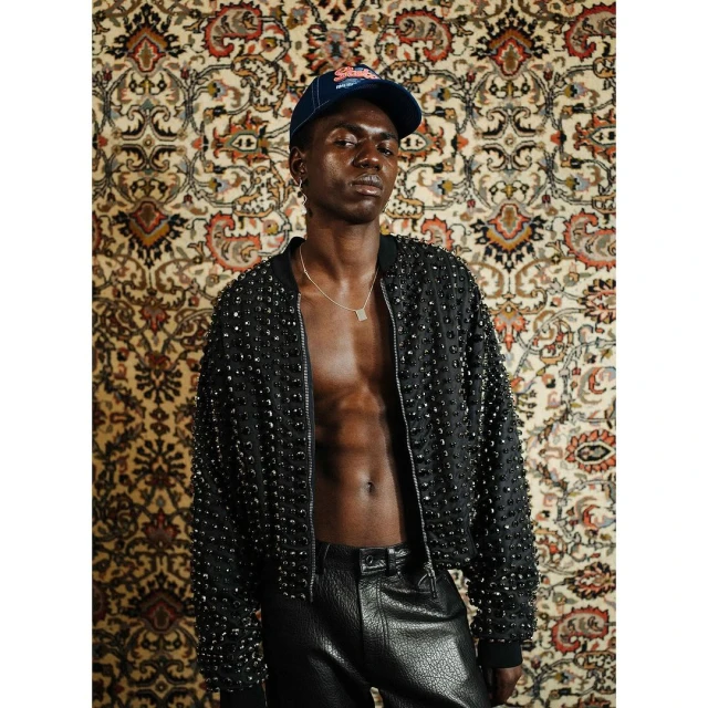 An African male model poses in black studded jacket and leather pants, accessorized with a necklace and baseball cap, against patterned wallpaper.