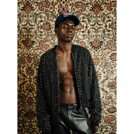 An African male model poses in black studded jacket and leather pants, accessorized with a necklace and baseball cap, against patterned wallpaper.