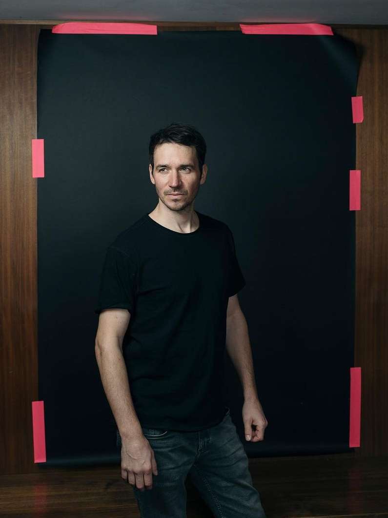 Portrait of a man in a black t-shirt posing in a studio with a black backdrop, lit by studio lighting.
