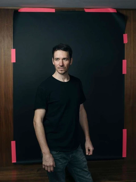 Portrait of a man in a black t-shirt posing in a studio with a black backdrop, lit by studio lighting.
