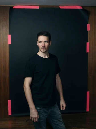 Portrait of a man in a black t-shirt posing in a studio with a black backdrop, lit by studio lighting.