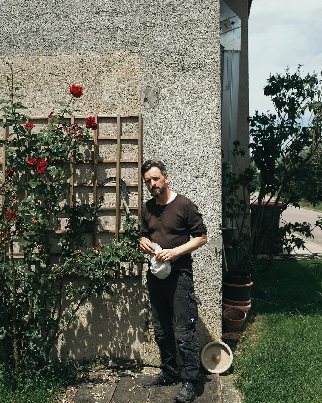 Man stands near a climbing rose on a sunny day in a backyard. He is holding a cap and wearing work clothes, with greenery and a building behind.