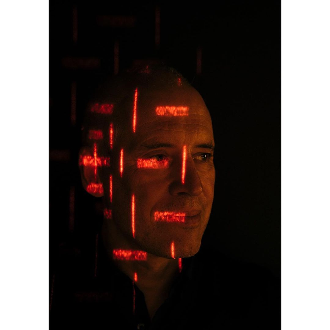 Portrait of an adult man with fair skin and a bald head, with red digital grid patterns overlayed on his face in a studio setting.