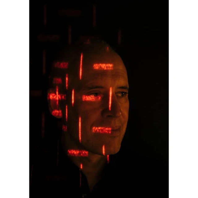 Portrait of an adult man with fair skin and a bald head, with red digital grid patterns overlayed on his face in a studio setting.