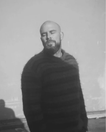 Monochrome portrait of a bald man with a beard wearing a dark fuzzy striped sweater against a plain background