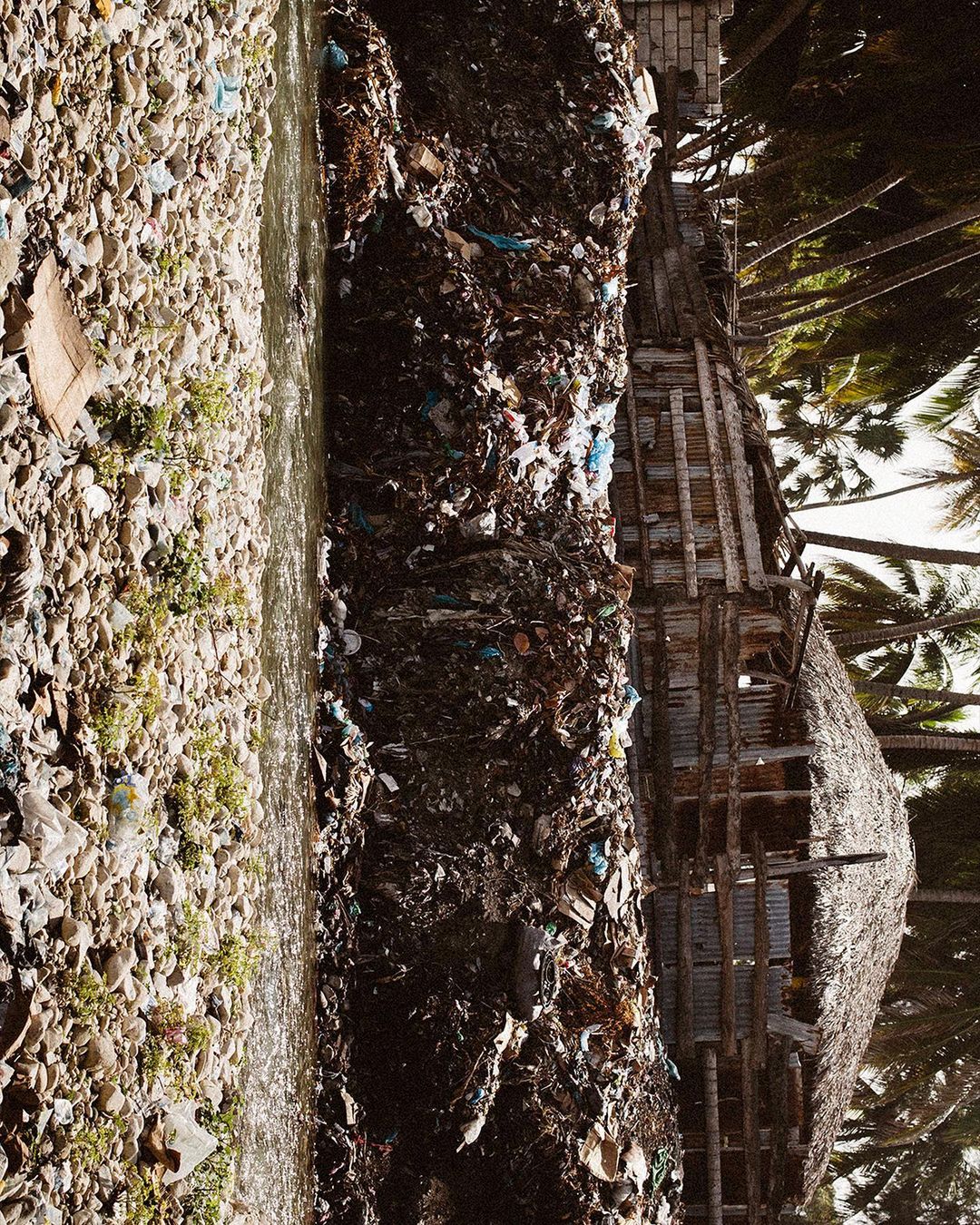 Polluted river with accumulated garbage flows next to a weathered wooden building and tropical trees, showcasing environmental degradation.