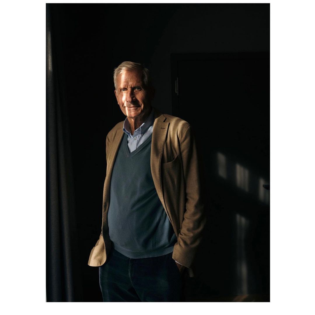 Elderly man in a blue sweater and khaki jacket stands in a dimly lit room with sunlight on his face, looking toward the camera.