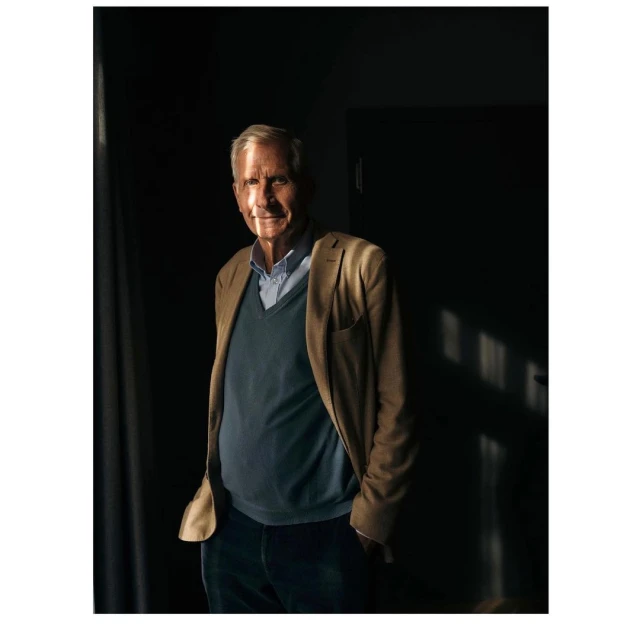 Elderly man in a blue sweater and khaki jacket stands in a dimly lit room with sunlight on his face, looking toward the camera.