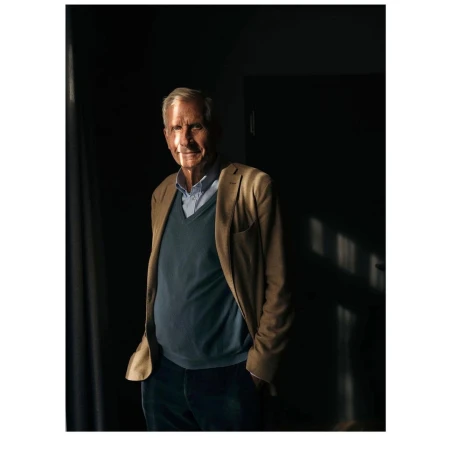 Elderly man in a blue sweater and khaki jacket stands in a dimly lit room with sunlight on his face, looking toward the camera.