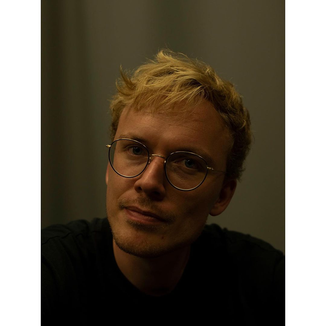 Close-up portrait of an adult male with blonde hair wearing round, thin-rimmed glasses, looking directly at the camera with a slight smile.