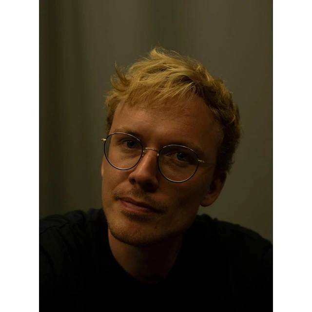 Close-up portrait of an adult male with blonde hair wearing round, thin-rimmed glasses, looking directly at the camera with a slight smile.