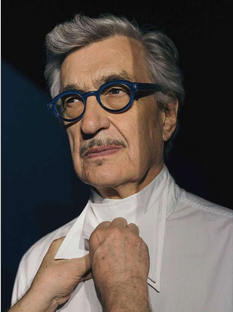 Portrait of an elderly man with grey hair and mustache, wearing blue glasses and a white shirt, photographed indoors against a dark background.