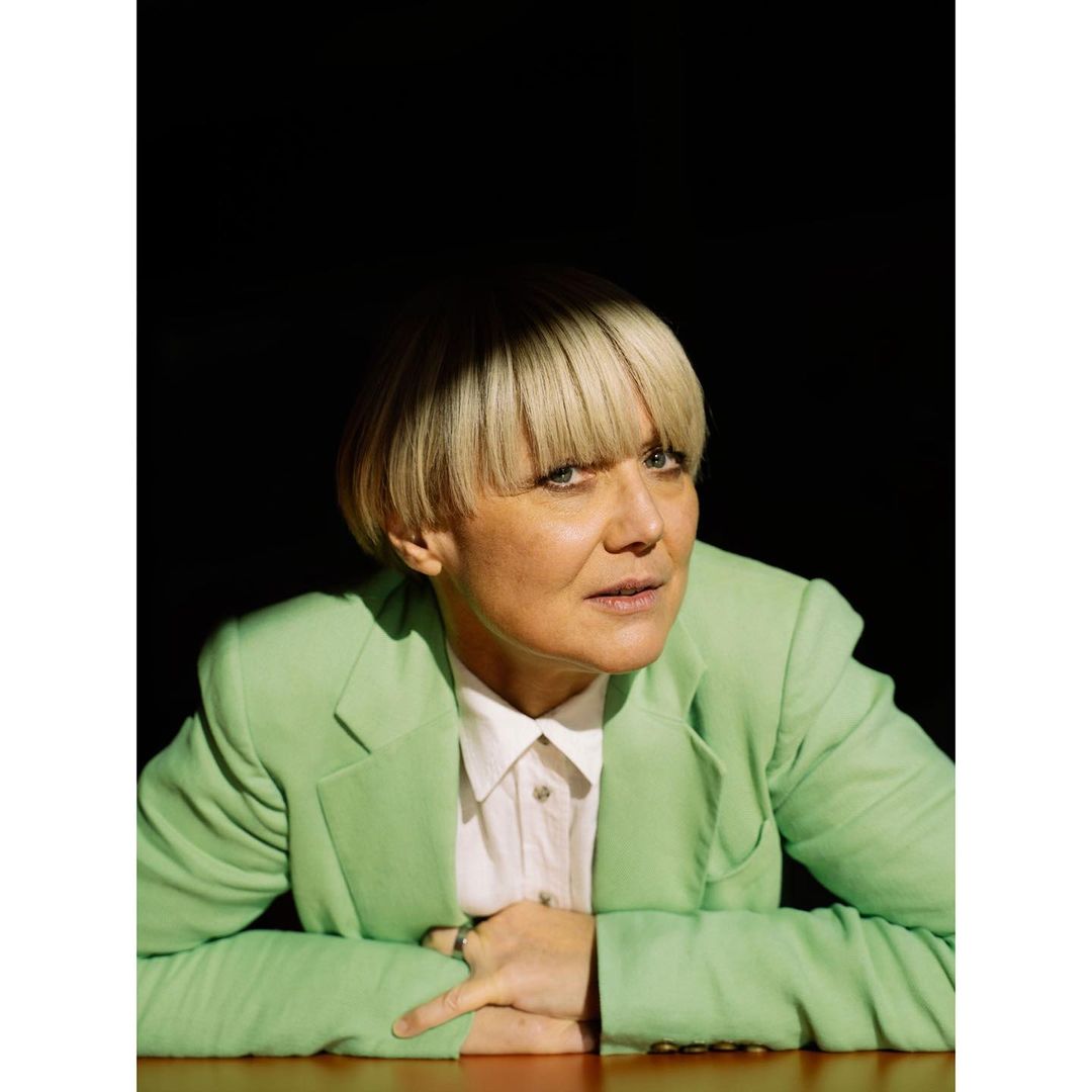 Blonde woman in a light green blazer poses indoors with a neutral expression, lit by diffused light and set against a dark background.