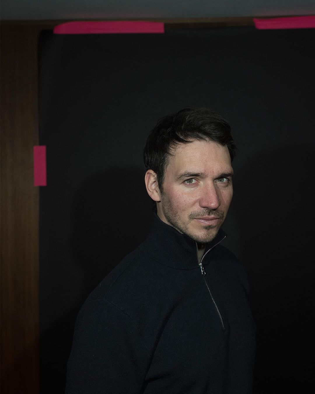 Headshot of an adult male with short dark hair posing in front of a dark background with pink tape, wearing a black zip-up sweater.