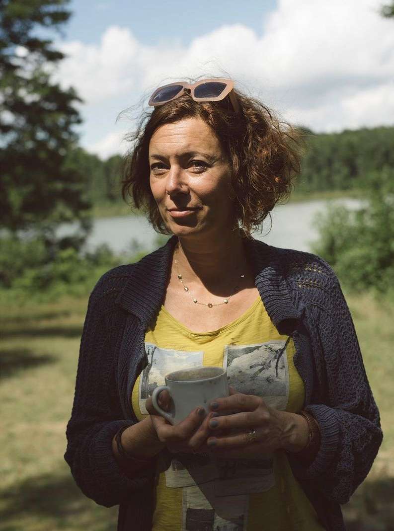 Adult woman holds a coffee mug outdoors in a green wooded area with a lake under a sunny sky during the daytime.