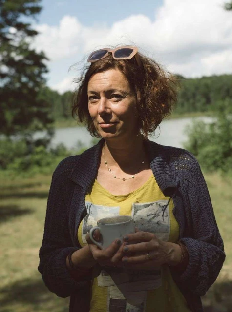 Adult woman holds a coffee mug outdoors in a green wooded area with a lake under a sunny sky during the daytime.