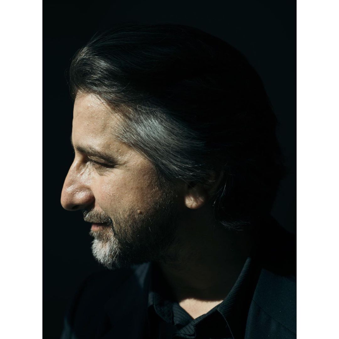Profile portrait of a mature adult male with grey and dark hair, a beard, and wearing a dark shirt, set against a dark background.
