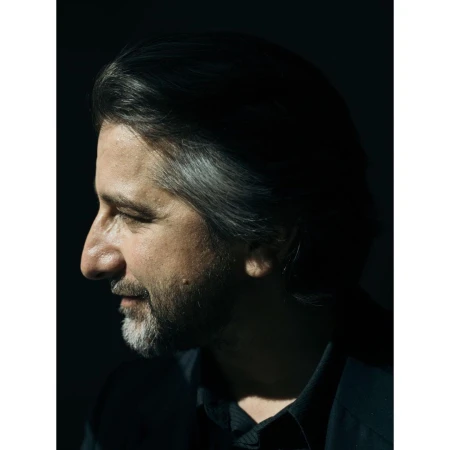 Profile portrait of a mature adult male with grey and dark hair, a beard, and wearing a dark shirt, set against a dark background.