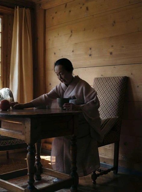 Adult woman wearing a kimono sits at a wooden table indoors, holding a cup in her hands near a window with a white curtain.