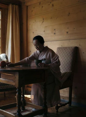 Adult woman wearing a kimono sits at a wooden table indoors, holding a cup in her hands near a window with a white curtain.