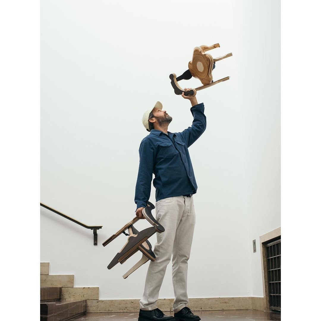 Man holding a wooden rocking chair above his head, with two more chairs in his other hand, standing on a staircase, indoors.
