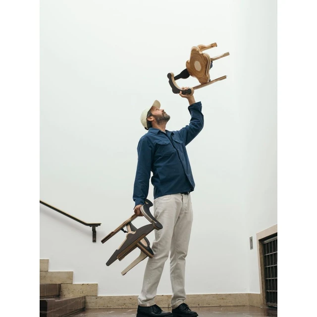 Man holding a wooden rocking chair above his head, with two more chairs in his other hand, standing on a staircase, indoors.