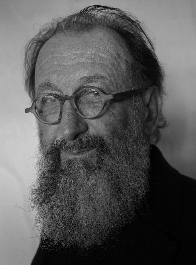 Black and white portrait of a mature man with a full beard, wearing glasses, and dressed in dark clothing against a plain background.