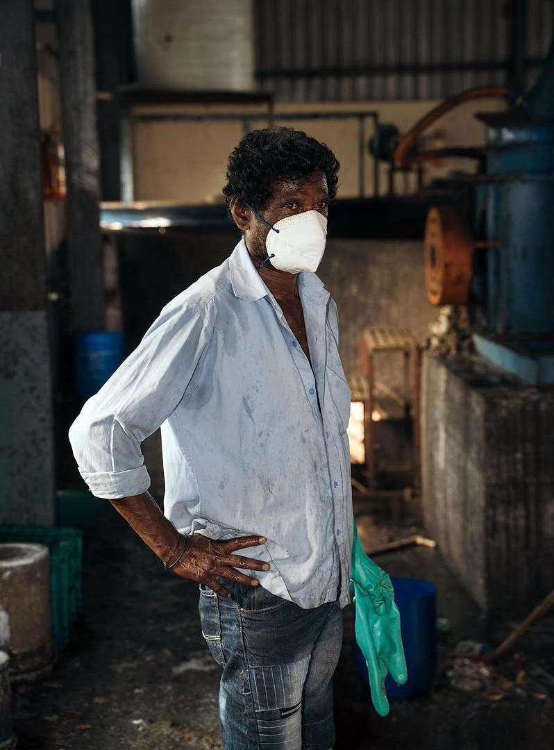Worker wearing a face mask and gloves in a factory setting with industrial machinery, wearing a blue button-down shirt and jeans.
