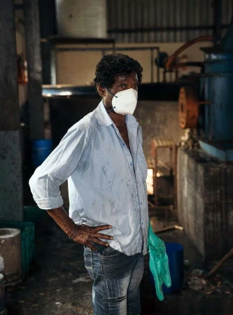 Worker wearing a face mask and gloves in a factory setting with industrial machinery, wearing a blue button-down shirt and jeans.