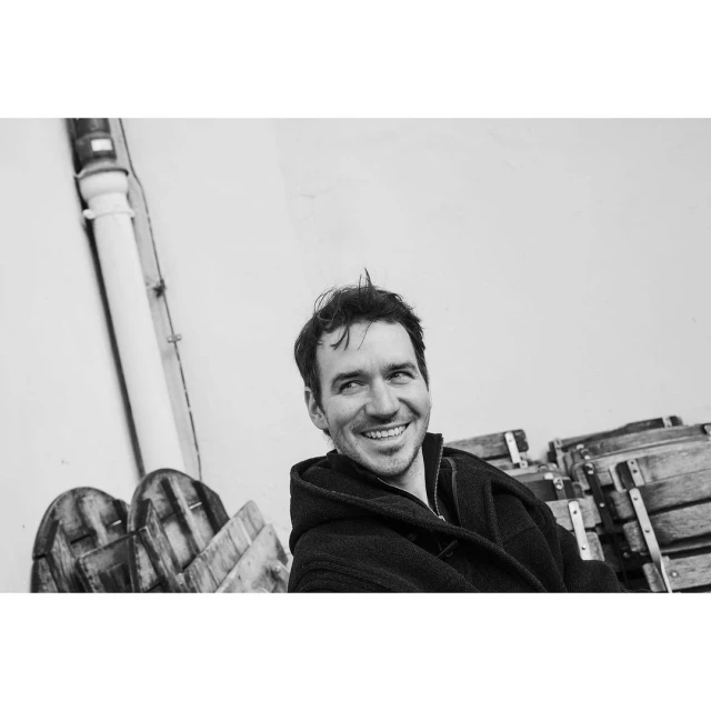 Monochromatic portrait of a smiling adult man wearing a dark hooded jacket, standing outdoors near wooden barrels and a light wall.